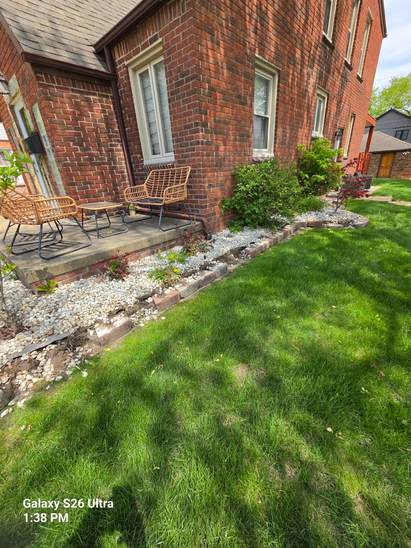 Side flower bed at brick home with weedy white stone and bare dirt patches before mulch refresh